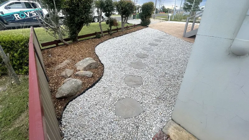 Curved gravel path with stepping stones meandering beside a building, bordered by mulch and small trees.