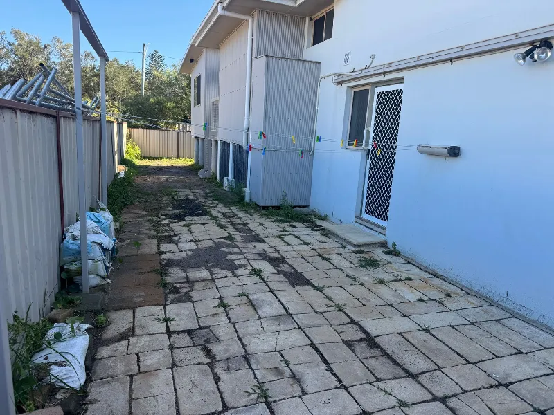 Narrow paved alley beside a blue house, with a clothesline, trash bags on the left, and a gate at the far end.