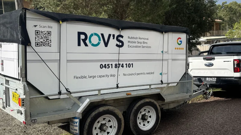 Trailered mobile skip-bin with branding 'ROV'S Rubbish Removal Mobile Skip Bin Excavation Services' hooked to a white pickup truck, roadside