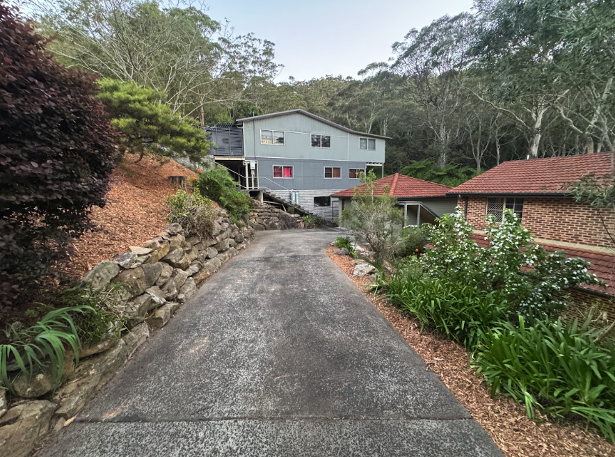 Paved driveway curves uphill toward a light-blue two-story house, with a stone retaining wall on the left and a brick house with a tiled roof on the right, surrounded by greenery.