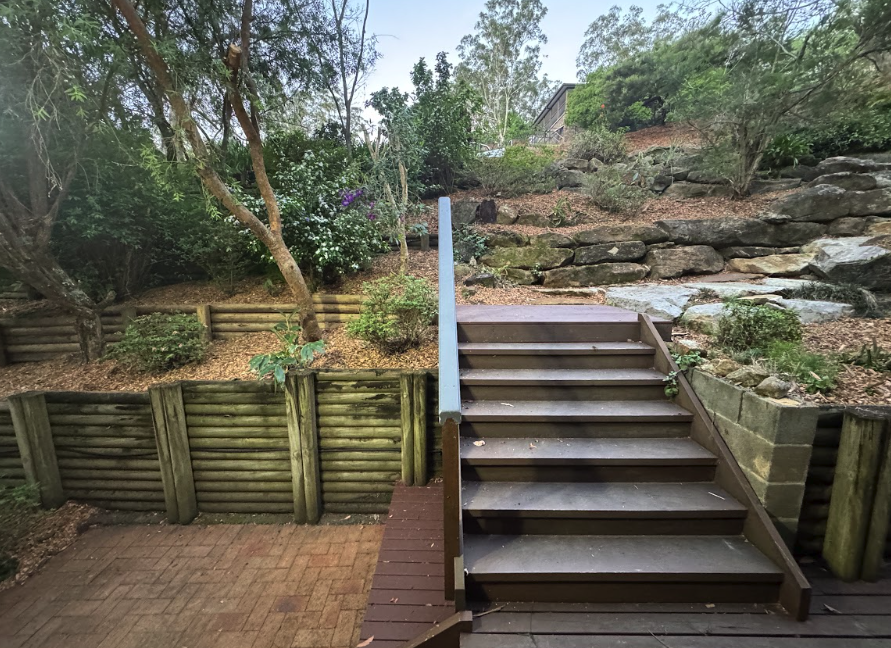Wooden deck with a wide brown staircase leading up to a terraced garden, stone retaining walls, and trees in the background.