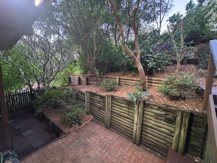 Terraced backyard garden with wooden retaining walls, trees, and shrubs on a mulched slope above a brick patio area on the ground.